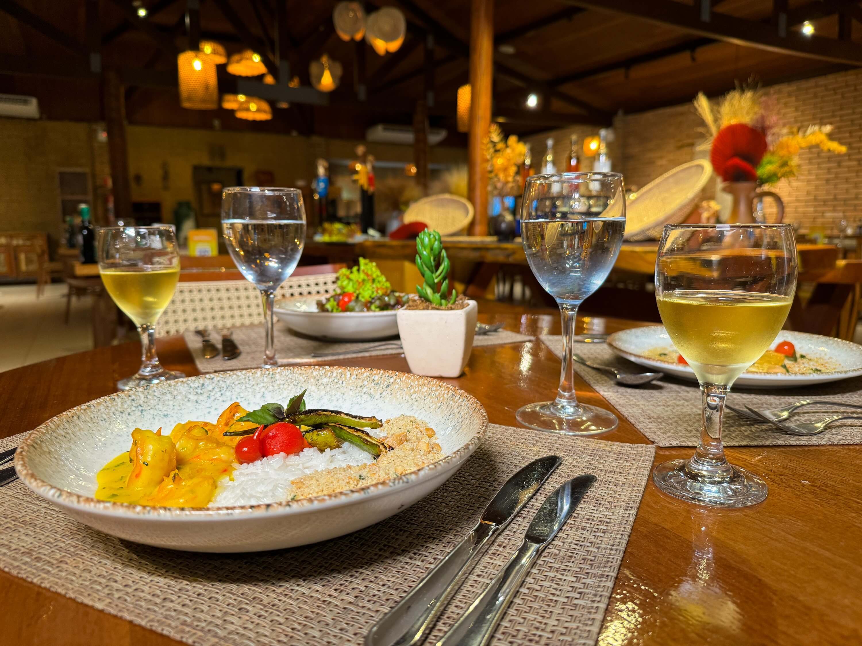 Mesa de jantar do Restaurante Mandacaru, com pratos de moqueca de camarão com arroz branco e farofa, acompanhados de vinho branco e água.